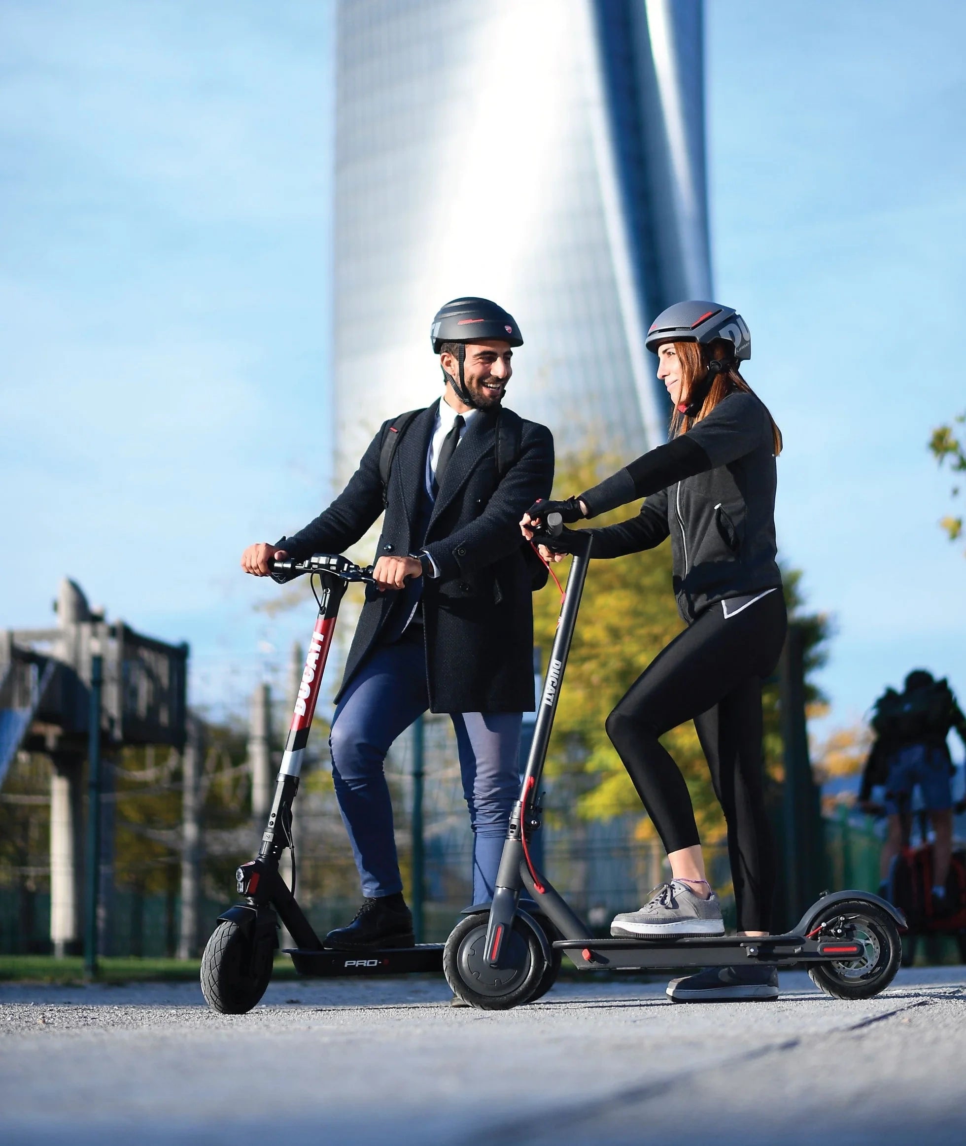 Two people riding electric scooters on a path with a fountain in the background