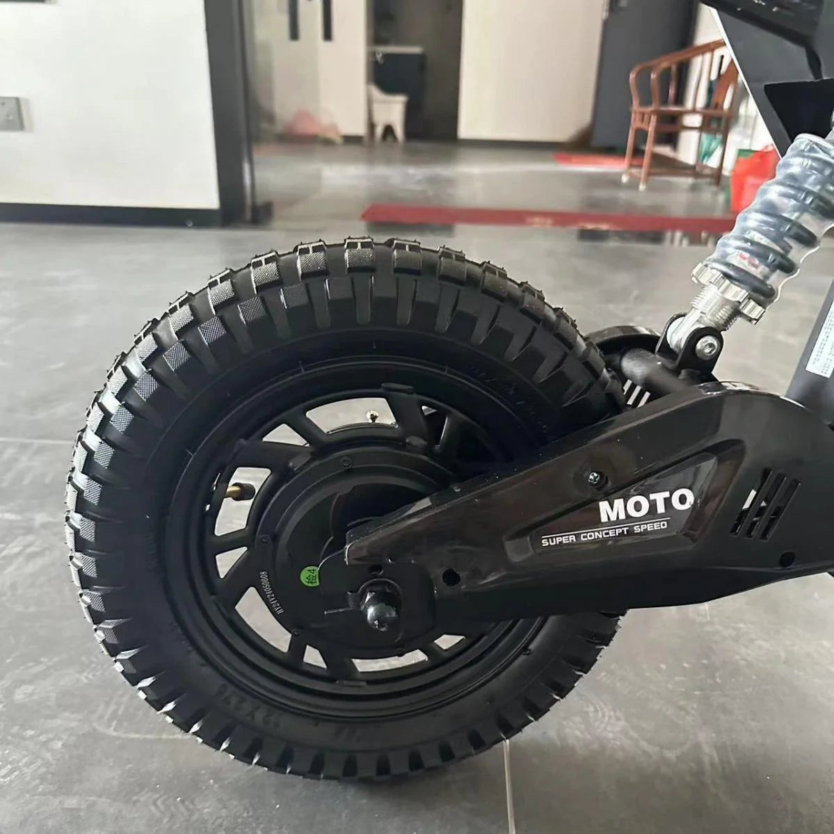 Close-up of a black electric scooter wheel with 'Moto' branding on a blurred indoor background.