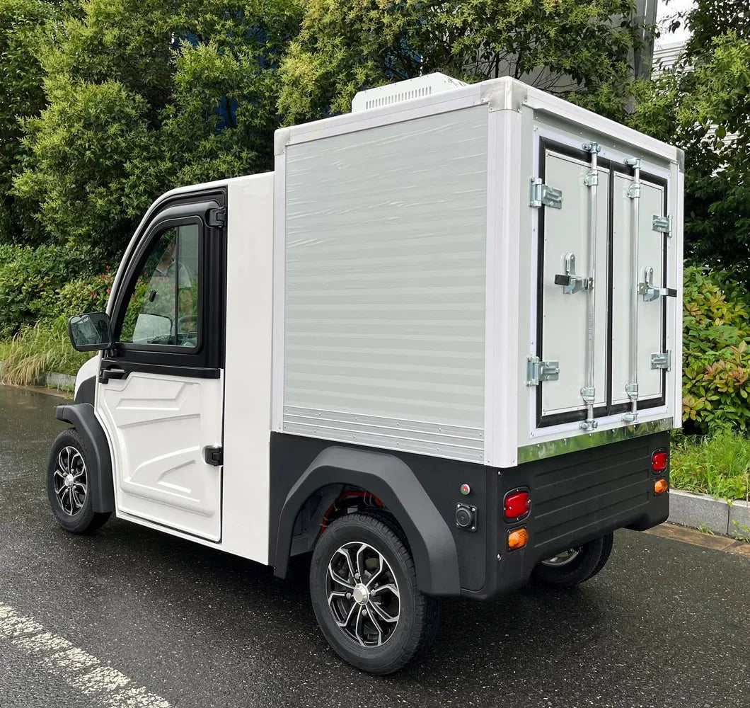 ocean cartz rear three-quarter view of a white electric refrigerated delivery vehicle with an enclosed cab, parked on a wet asphalt road. This compact electric NEV or refrigerator work cart features a silver insulated rear cargo box with double doors and a top-mounted refrigeration unit. The vehicle has black trim, black fender flares, and custom black and silver wheels, highlighting its use for cold chain last-mile delivery.