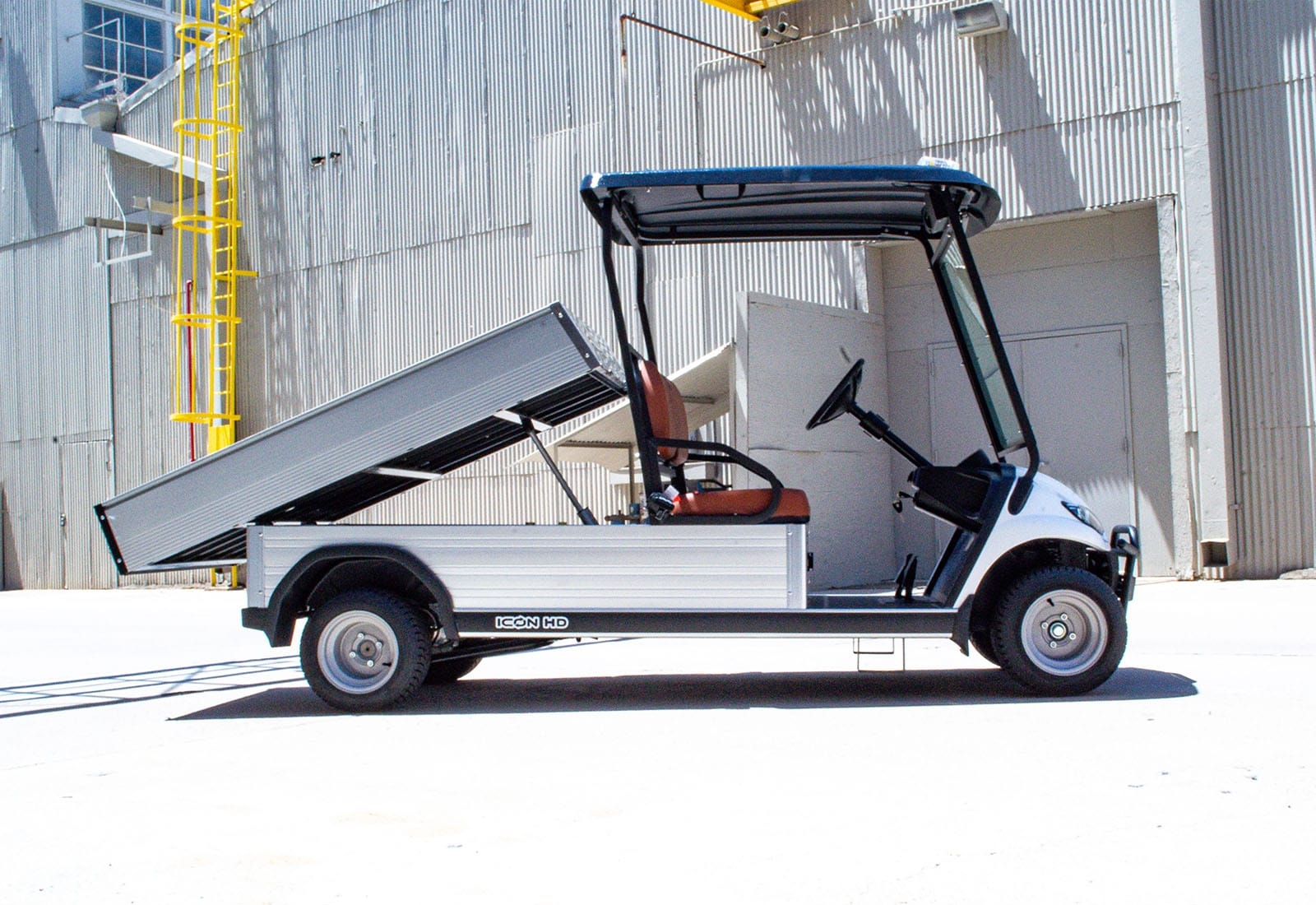 ocean cartz side profile view of a white utility electric vehicle with a fully raised silver aluminum dump bed, demonstrating the hydraulic function. This specialized electric work cart features a black roof and a single row of brown seating. The dump cart is parked in front of a corrugated metal industrial building, highlighting its use as a heavy-duty hauling utility vehicle.