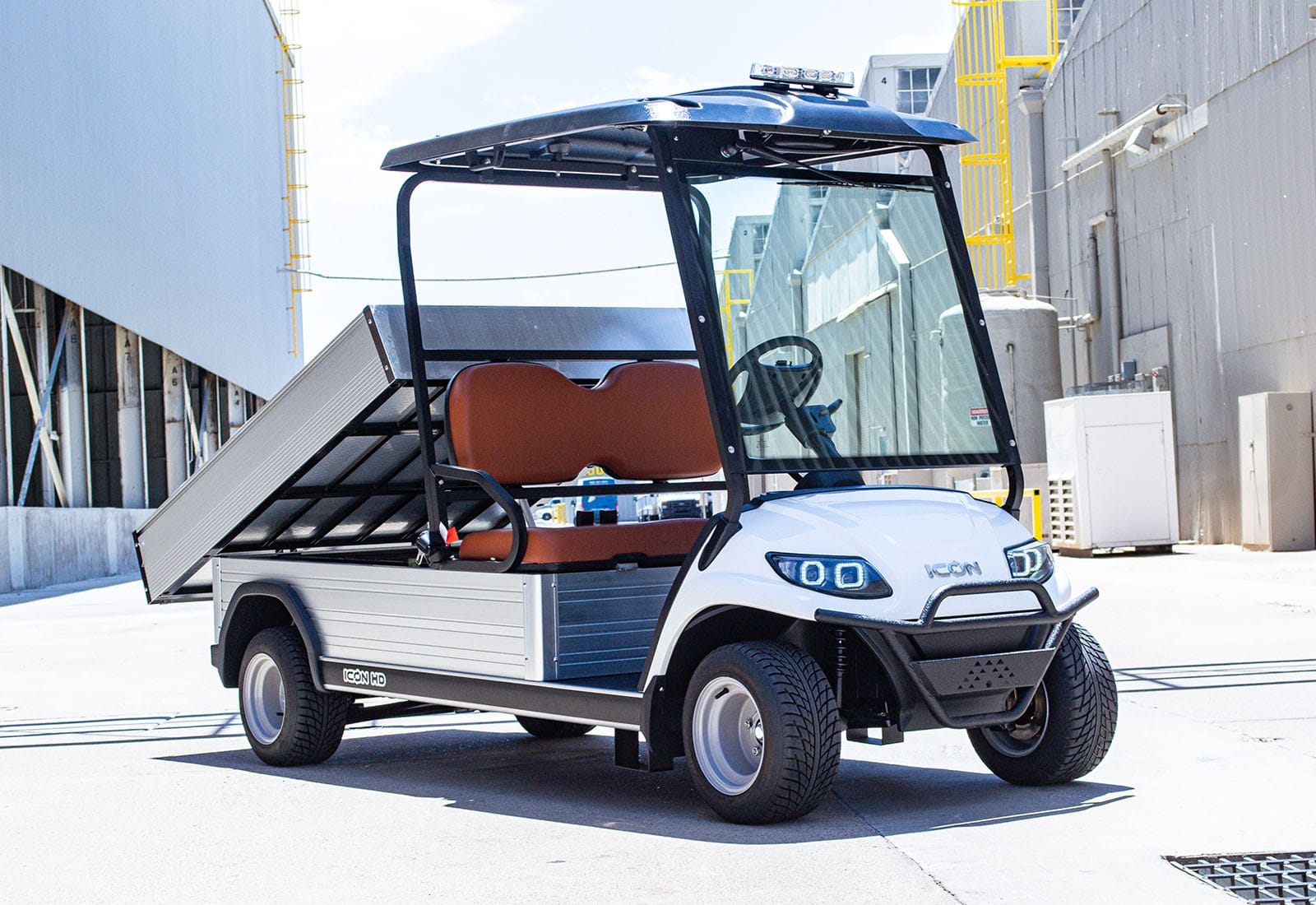 ocean cartz three-quarter front view of a white utility electric vehicle with a fully electric dump bed raised for unloading, parked in an industrial area. This specialized electric work cart features a black roof, a black brush guard, and brown two-seater upholstery. The utility golf cart has custom wheels and a clear windshield, with the large aluminum dump bed prominently displayed.