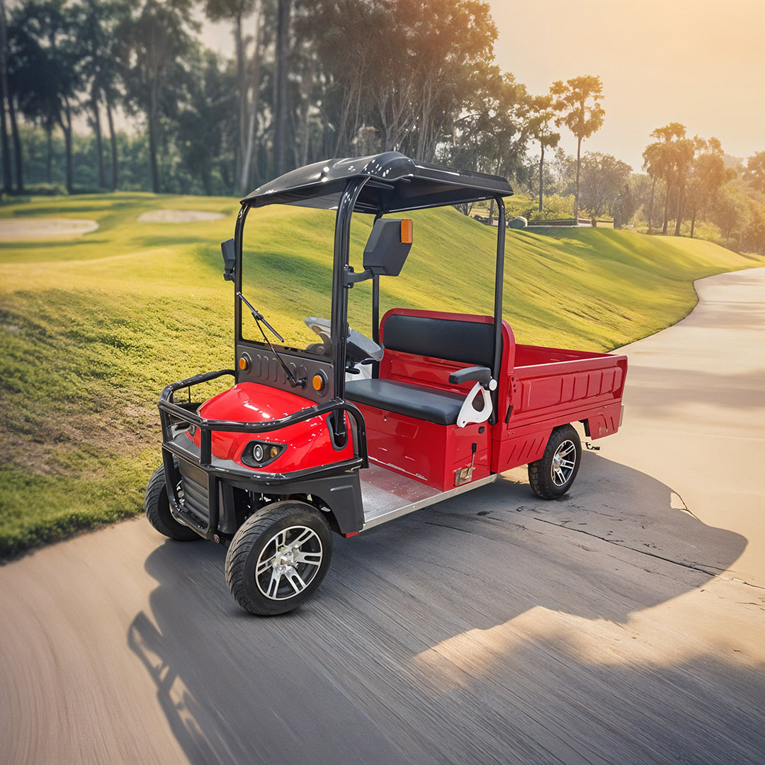 ocean cartz three-quarter front view of a red utility electric vehicle with a flatbed and a black roof, driving on a paved cart path. This two-seater electric work cart features black seats, a black front brush guard, and custom silver alloy wheels. The electric utility truck is pictured in a sunny, expansive outdoor setting, likely a resort or golf course, with green hills and tall trees in the background.