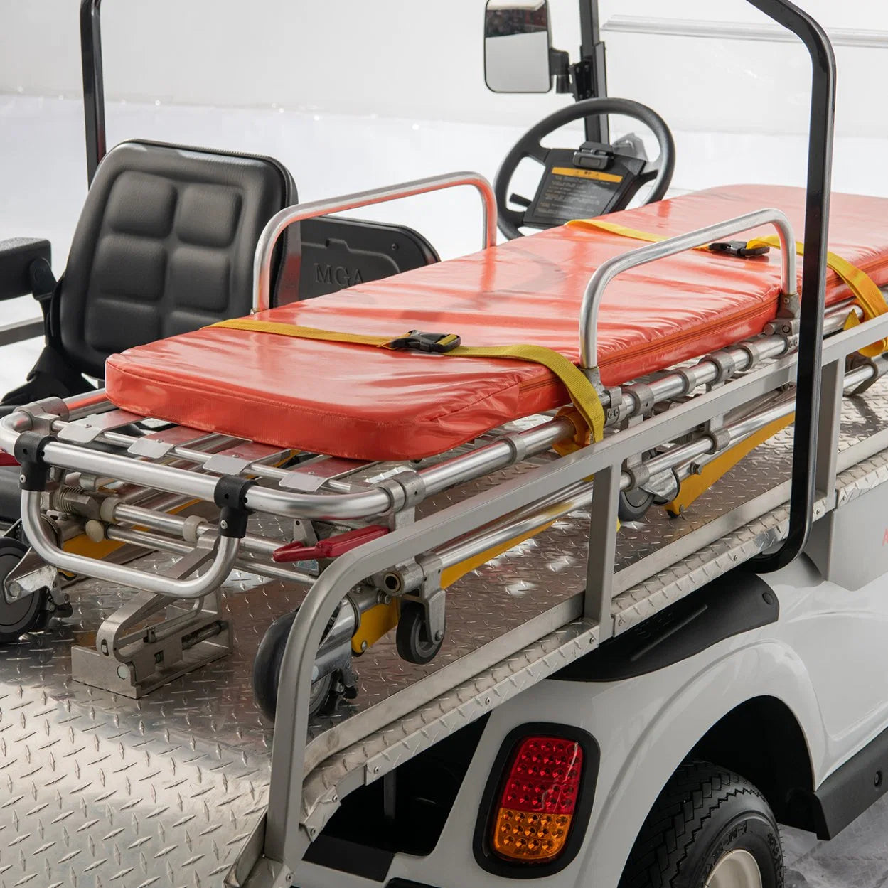ocean cartz close-up interior view of a white ambulance golf cart focusing on the patient transport area. The image clearly shows a metallic, diamond-plated rear deck holding a silver emergency stretcher with an orange pad and yellow safety straps. The driver's black seat and steering wheel are visible in the background, highlighting the specialized equipment of this rescue golf cart or medical utility vehicle.