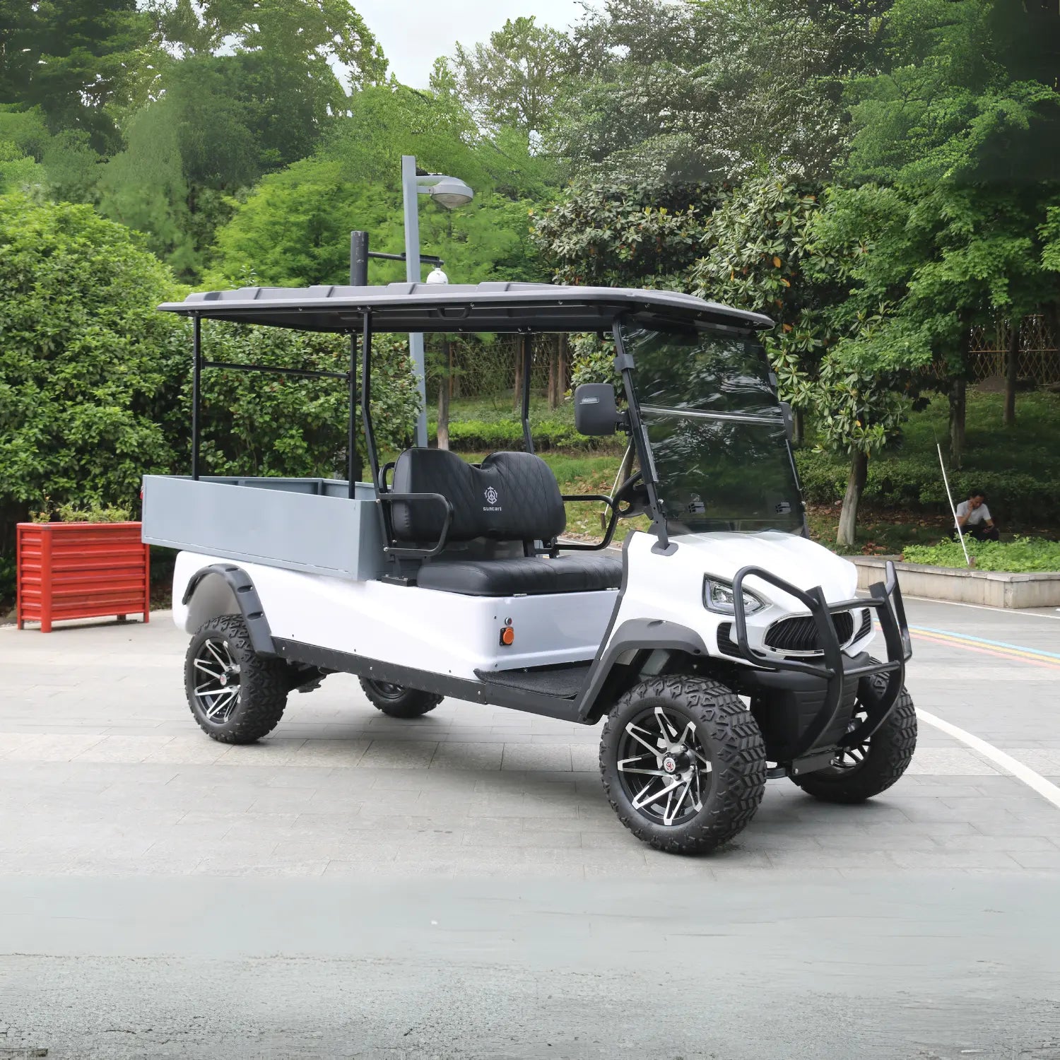 ocean cartz side view of a white utility golf cart with a large, grey rear cargo box and a black roof, parked on an asphalt path. This lifted electric vehicle is an off-road utility cart featuring black quilted seats, a front brush guard, and aggressive all-terrain tires. The wheels are custom black and silver alloy designs, and the vehicle is shown in a park or campus setting surrounded by dense green trees.