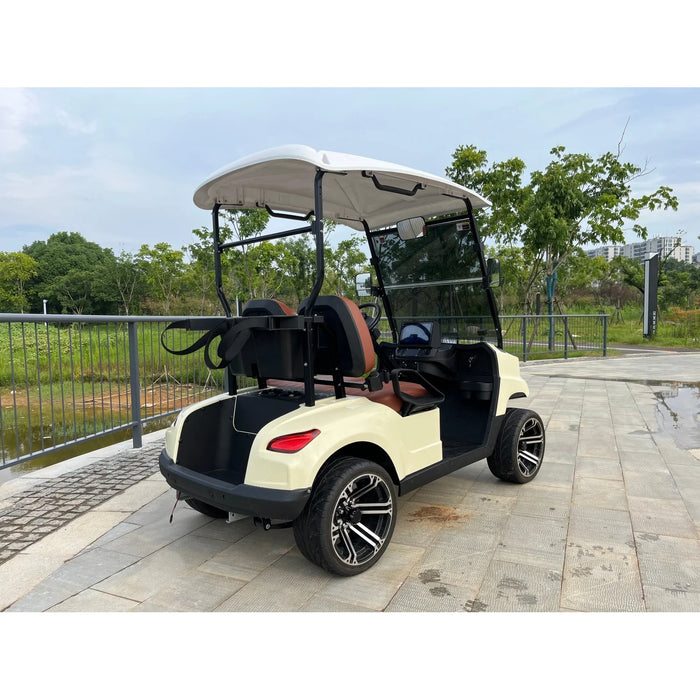 ocean cartz rear three-quarter view of a cream-colored four-seater luxury golf cart with a white roof, parked on a paved path overlooking a body of water in a park setting. This electric vehicle features brown and black seats, a rear cargo area with a bag attachment, and a tinted windshield. The cart is equipped with low-profile tires and custom black and silver alloy wheels, highlighting its sleek NEV or resort transport design.