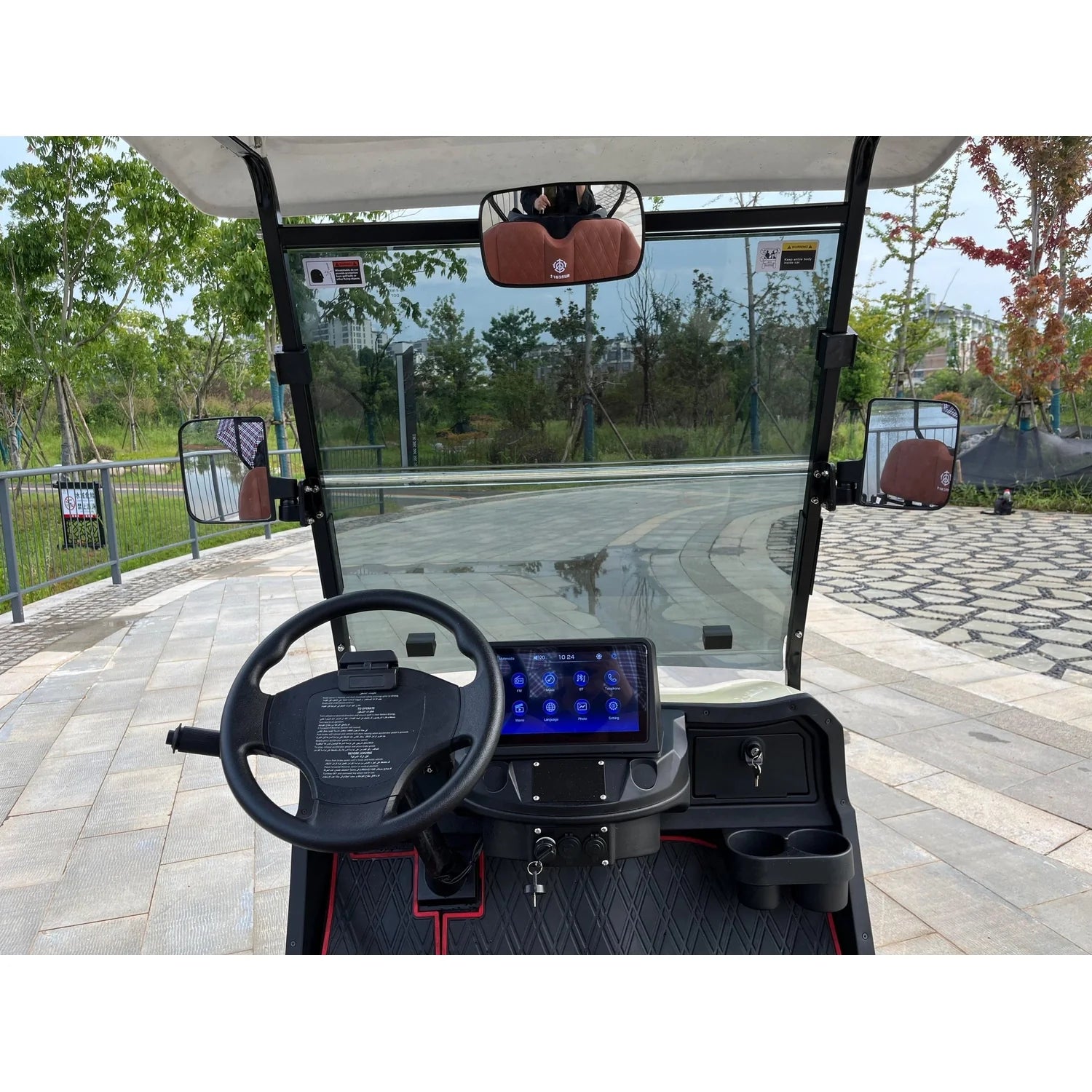 ocean cartz close-up interior view of the driver's cabin in a cream-colored luxury golf cart. The image focuses on the modern, black dashboard, which includes a centered digital touchscreen display, a black steering wheel, and dual cup holders. The cabin features a panoramic windshield, side mirrors, and a rearview mirror with a brown accent. The electric vehicle is parked on a paved stone path in a green, park-like setting, highlighting the interior technology of this NEV.
