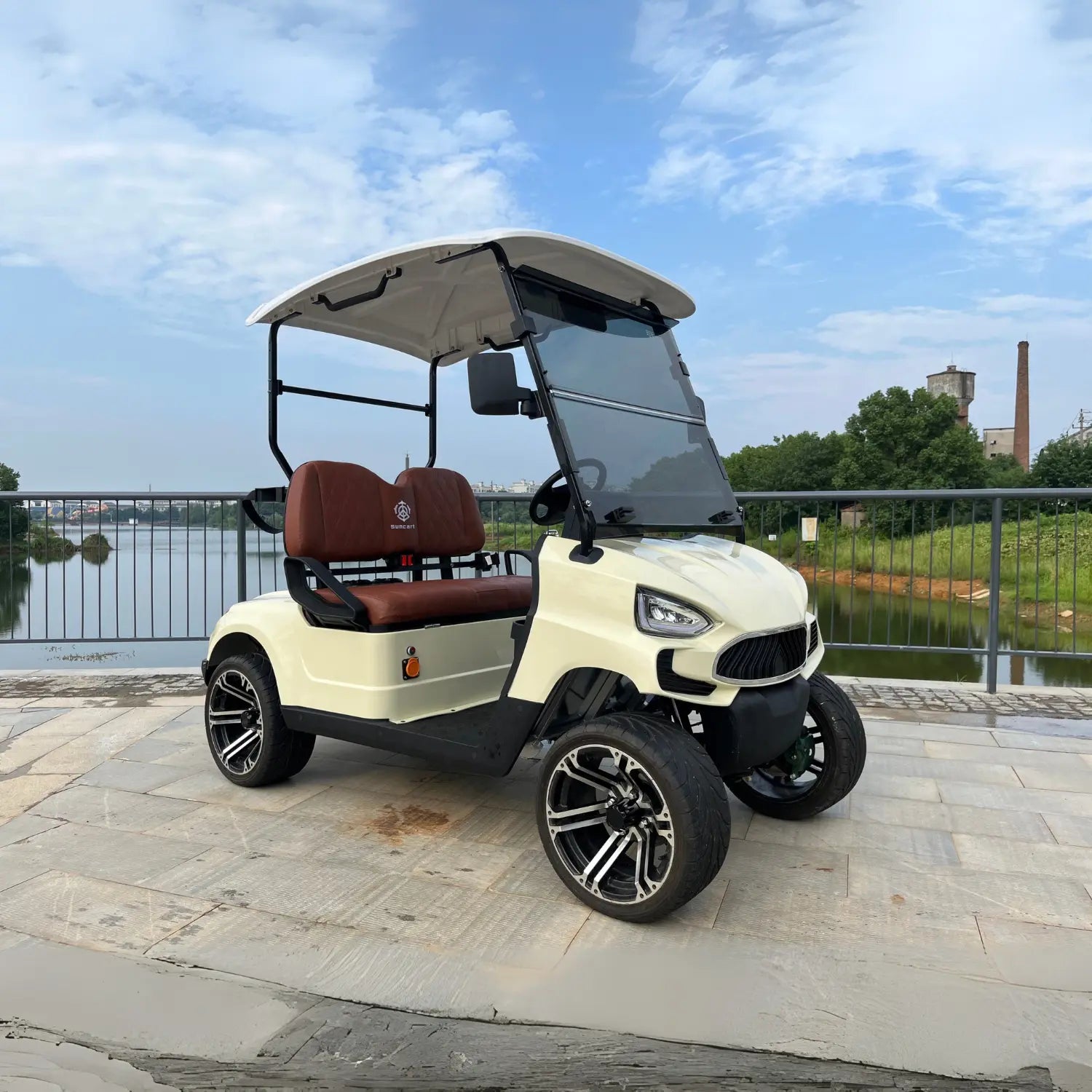 ocean cartz three-quarter front view of a cream-colored four-seater luxury golf cart with a white roof, parked on a paved walkway next to a body of water. This electric vehicle features brown leather seats with diamond stitching, a modern front end, and a tinted windshield. The lifted golf cart is equipped with all-terrain tires and custom black and silver alloy wheels, highlighting its stylish and premium resort or NEV design.