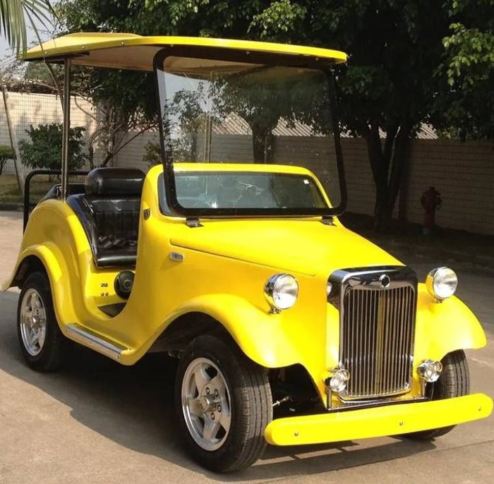 ocean cartz three-quarter front view of a bright yellow classic antique-style golf cart with a matching yellow roof. This two-seater vintage electric vehicle features a traditional chrome grille, round headlights, and a large windshield. The classic golf cart has black seats and is equipped with multi-spoke silver wheels, parked on asphalt in a sunny outdoor setting with trees and a white fence in the background.