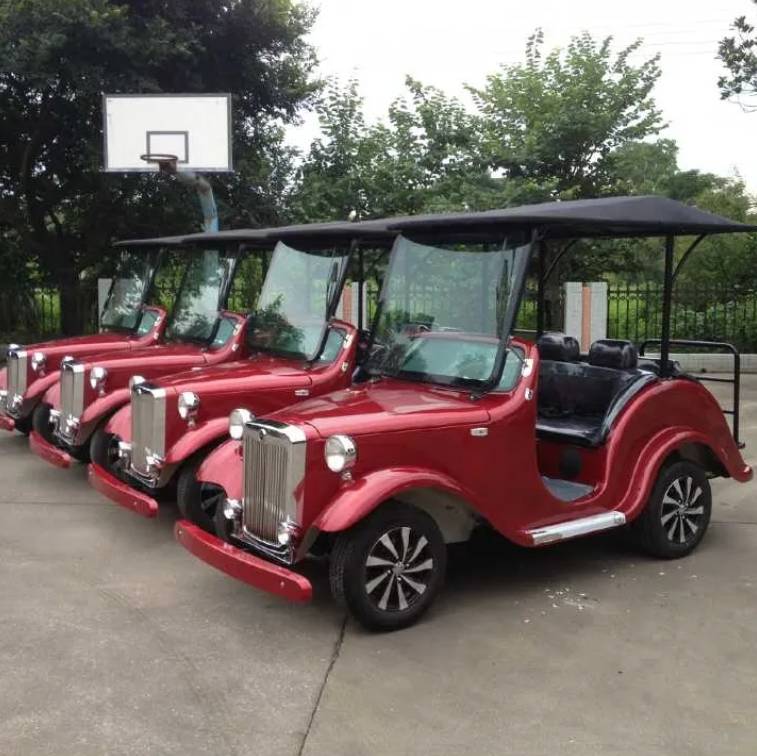 ocean cartz three-quarter front view of a line of three dark red classic antique-style golf carts with black roofs, parked on a paved court. The front vintage electric vehicle features a large chrome grille, round headlights, black seats, and custom black and white wheels. These multi-passenger classic carts are shown in an outdoor park setting with a basketball hoop and trees in the background, highlighting their use as resort transport vehicles.
