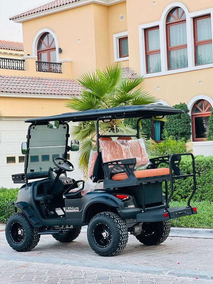 ocean cartz rear three-quarter view of a matte black off-road golf cart with large, knobby all-terrain tires and a rear facing seat. The lithium electric vehicle features orange-brown upholstery, a black roof, and black custom wheels. The lifted four-seater golf car is parked on a paver driveway in front of a luxurious, light-colored Mediterranean-style villa with arched windows and a palm tree.