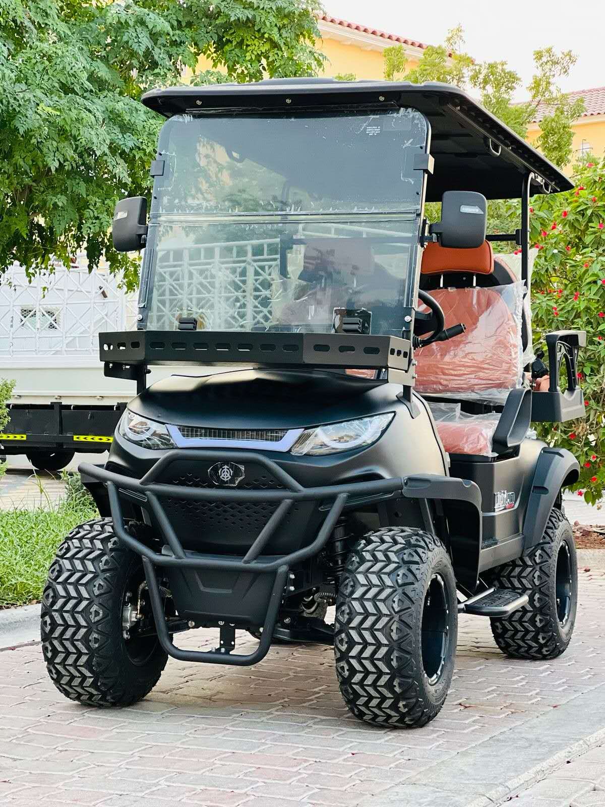 ocean cartz front view of a matte black off-road golf cart parked on a paved residential street. This lifted electric vehicle features a heavy-duty front brush guard, massive all-terrain tires on black wheels, and a front cargo basket. The four-seater utility cart has brown upholstered seats and is surrounded by lush green foliage and houses in the background.