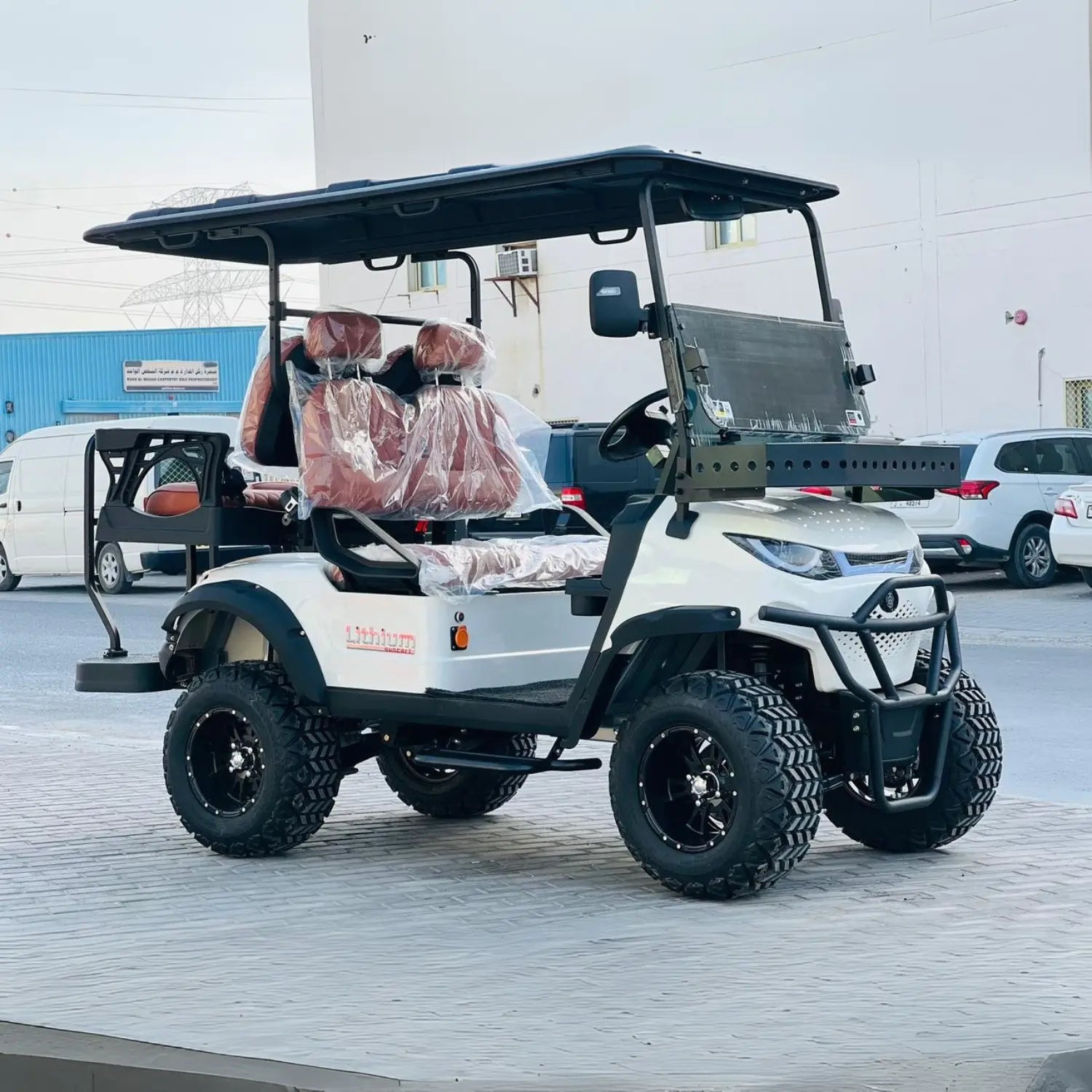 ocean cartz side view of a white lifted golf cart with a black roof and black trim. The off-road electric vehicle features large, aggressive all-terrain tires on black custom wheels and a front brush guard. It has brown leather seats with plastic coverings and a "Lithium" badge on the side. The four-seater utility cart is parked on a paved lot with industrial buildings and other cars in the background.