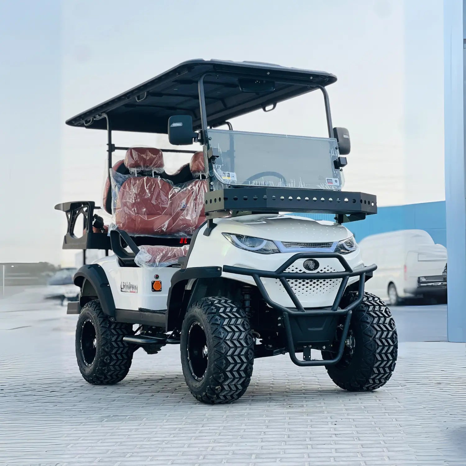 ocean cartz dramatic, low-angle front view of a white lifted golf cart with a black roof and black accents. This off-road electric vehicle features huge, aggressive all-terrain tires on black custom wheels, a heavy-duty brush guard, and a front cargo basket mounted above the hood. The four-seater utility cart has dark red or brown leather seats and is pictured outdoors in an open area with a bright sky.
