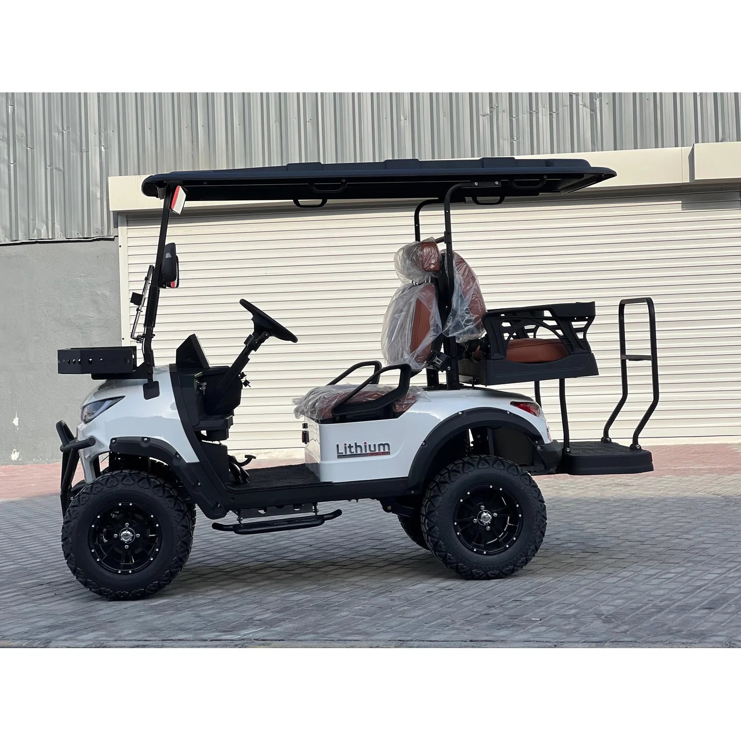 ocean cartz side profile view of a white lifted golf cart with a black roof and black fender flares. This lithium electric vehicle is an off-road four-seater utility cart featuring aggressive all-terrain tires on black custom wheels and a brown leather seat package. The cart has a front brush guard and a rear fold-down seat or cargo platform with safety rails. It is parked outside an industrial building with a large roll-up door.