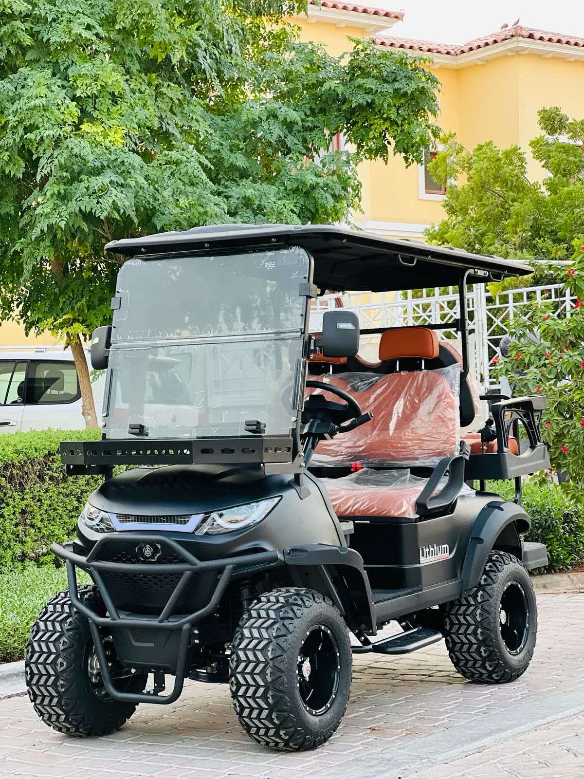 ocean cartz full view of a matte black off-road golf cart with aggressive styling and brown leather seats. The four-seater electric vehicle is equipped with a rugged brush guard, large, knobby all-terrain tires on black wheels, and a front cargo basket or rack. The lifted utility cart is parked on a paved residential street with lush green trees and a yellow stucco home in the background.