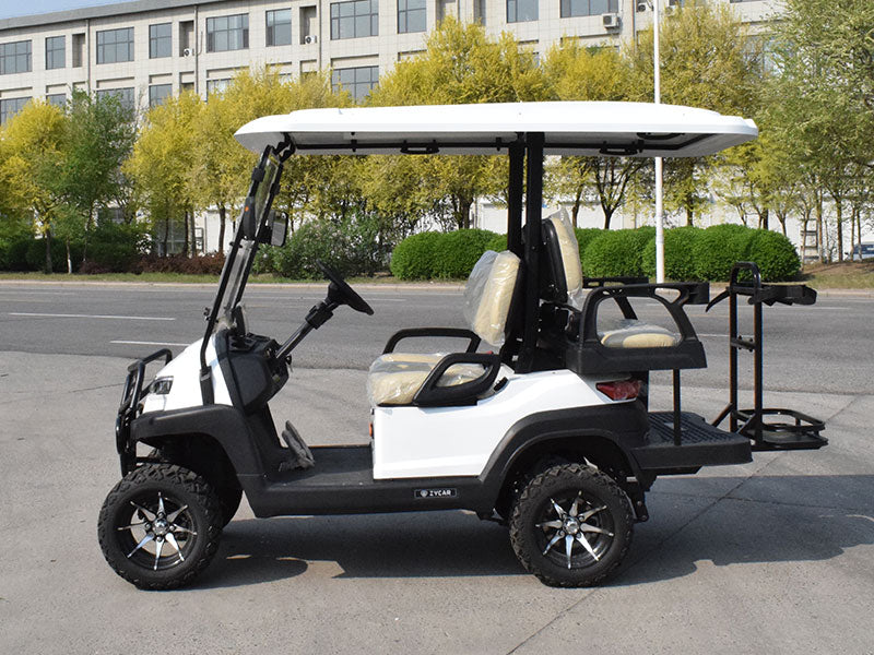 ocean cartz side profile view of a white six-seater golf cart with a white roof, parked on a street near a commercial building. This lifted off-road electric vehicle features a rear-facing flip seat, tan or cream-colored seats, and a cargo platform with a golf bag holder rack. The all-terrain golf cart is equipped with large, knobby off-road tires on custom black and silver wheels.