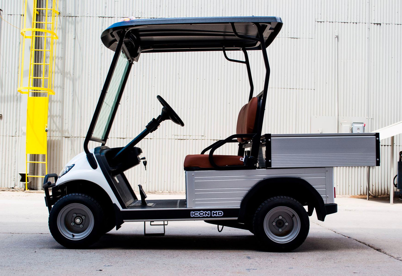 ocean cartz side profile view of a white utility electric vehicle with a black roof and a silver aluminum rear cargo bed with drop-down sides. This specialized two-seater electric work cart features brown seats, a steering wheel, and silver utility wheels. The electric utility golf cart is parked on a paved surface next to a corrugated metal industrial building, with a yellow ladder visible in the background, highlighting its heavy-duty property maintenance function.