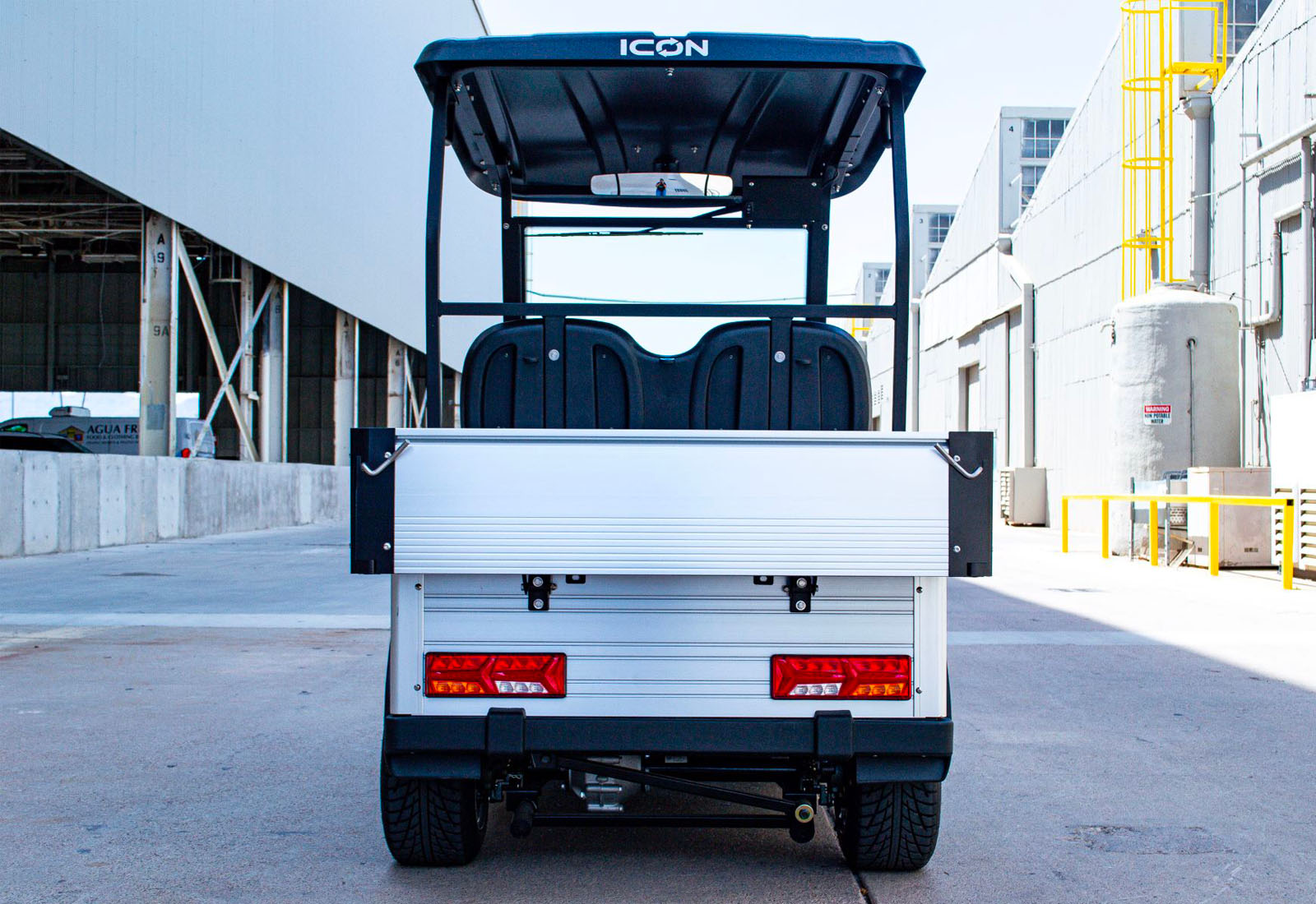 ocean cartz rear view of a white utility electric vehicle with a black roof and a silver aluminum rear cargo bed with the tailgate closed, parked in an industrial corridor. This specialized two-seater electric work cart features black bucket seats, rectangular red LED taillights, and black fender flares. The image highlights the heavy-duty rear hauling capacity and the overall rugged design of the electric utility golf cart.