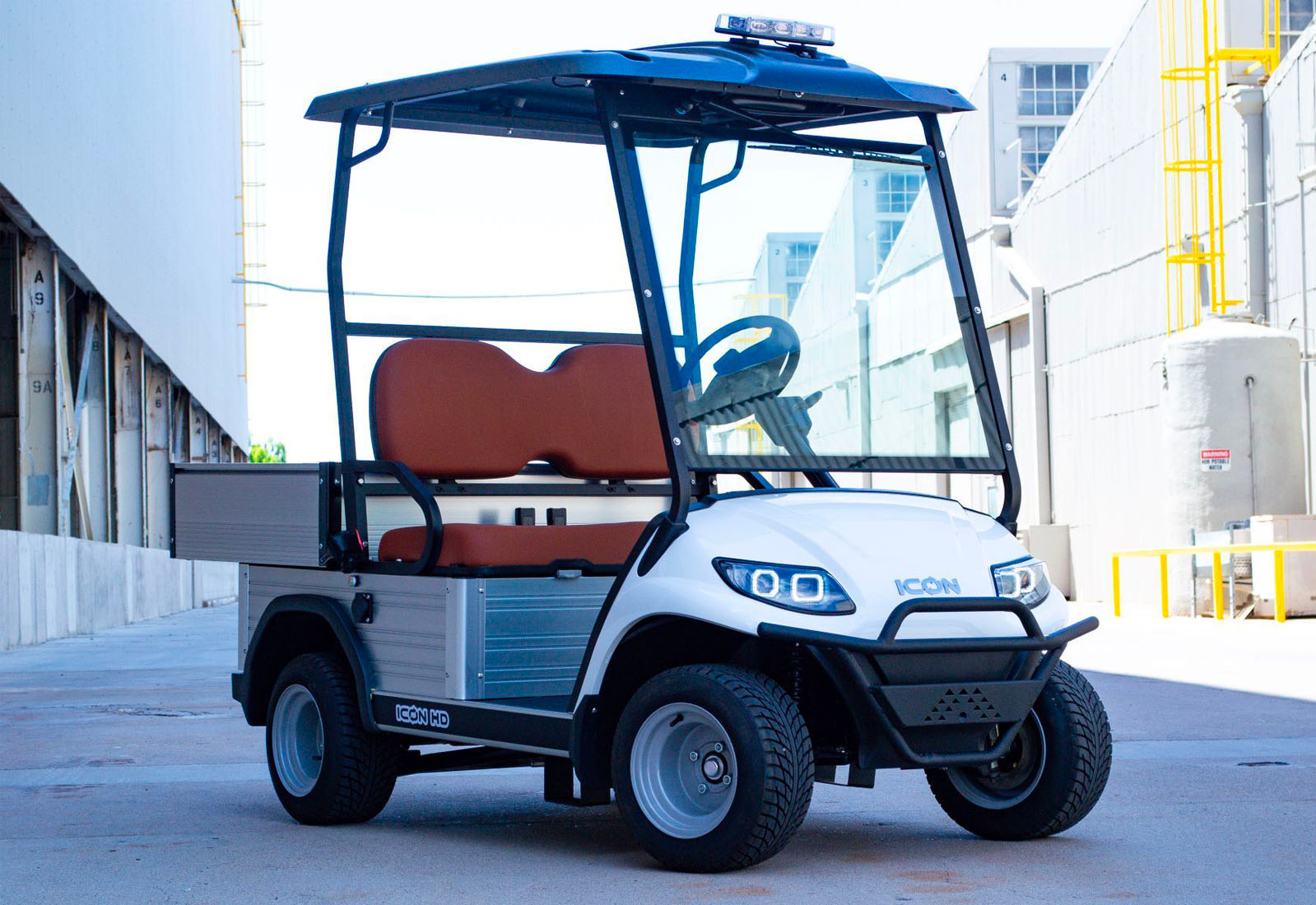 ocean cartz three-quarter front view of a white utility electric vehicle with a black roof and a silver aluminum rear cargo bed, parked in an industrial setting. This specialized two-seater work cart features brown seats, a protective front brush guard, a large windshield, and a light bar on the roof. The electric utility golf cart is equipped with silver utility wheels and all-terrain tires, highlighting its heavy-duty hauling and property maintenance function.