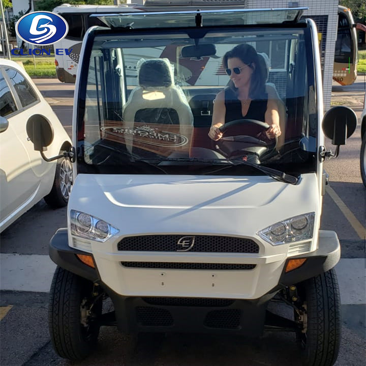 ocean cartz head-on view of a white two-seater electric vehicle with a fully enclosed cab. A woman wearing sunglasses is sitting in the driver's seat. This compact NEV or low-speed electric car features a modern front end, a small grille, and a clear windshield. The image is taken outdoors in a parking lot, with other vehicles and a bus visible in the background, showing the electric vehicle in use.