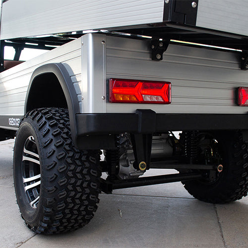 ocean cartz close-up rear view of the silver aluminum cargo bed and the off-road tires on a lifted electric utility vehicle. This shot highlights the robust rear suspension, the black and silver custom wheel, and the large, knobby all-terrain tire. The rear panel of the cargo bed features a modern, rectangular LED taillight, emphasizing the heavy-duty and rugged design of the electric work cart.