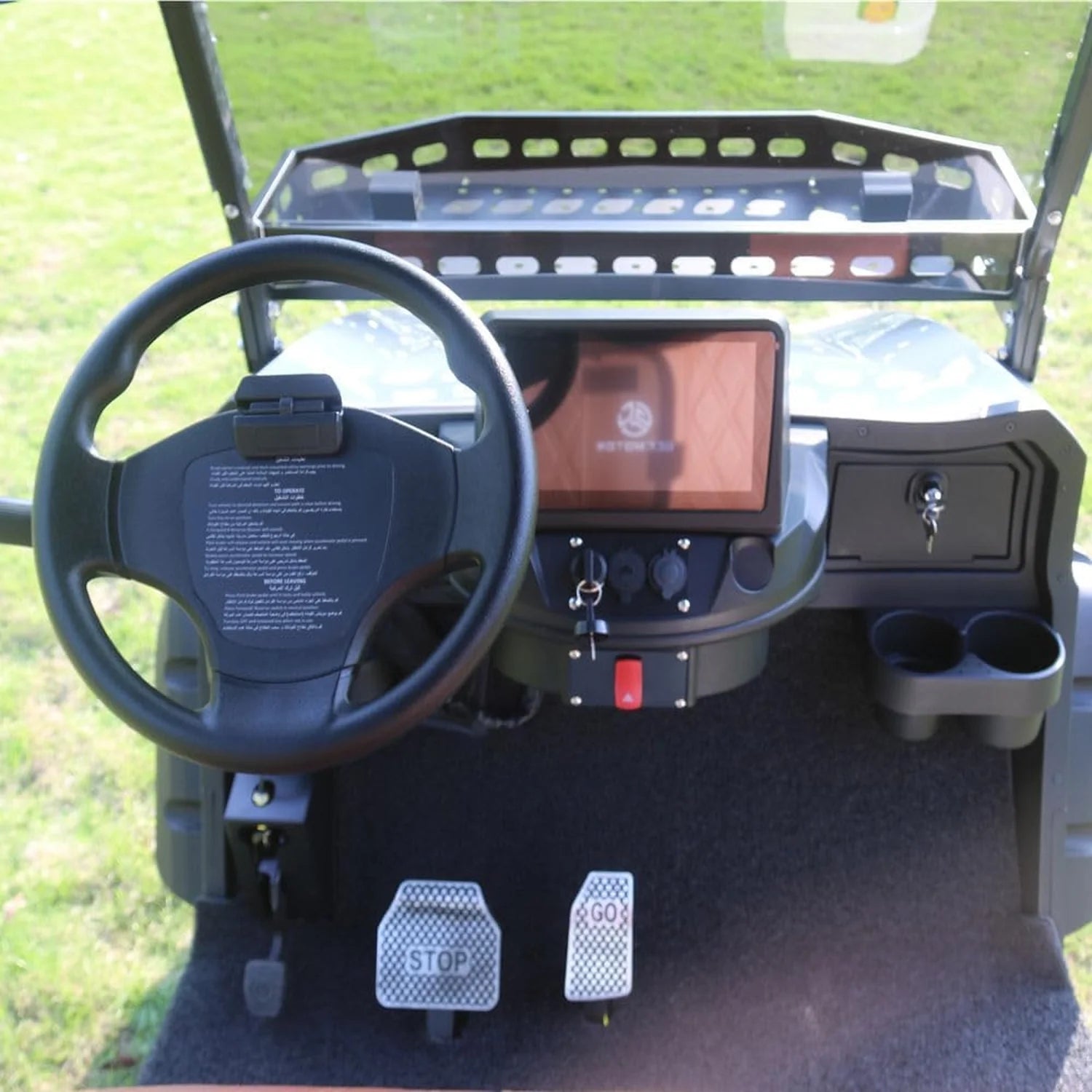 ocean cartz close-up interior view of the dashboard and controls of a luxury golf cart. The image highlights the digital touchscreen display, a black three-spoke steering wheel, and the driver's floor area with metallic "STOP" and "GO" pedal covers. The dashboard includes the ignition switch, accessory power ports, and two cup holders. This detailed shot emphasizes the modern features of the electric vehicle.