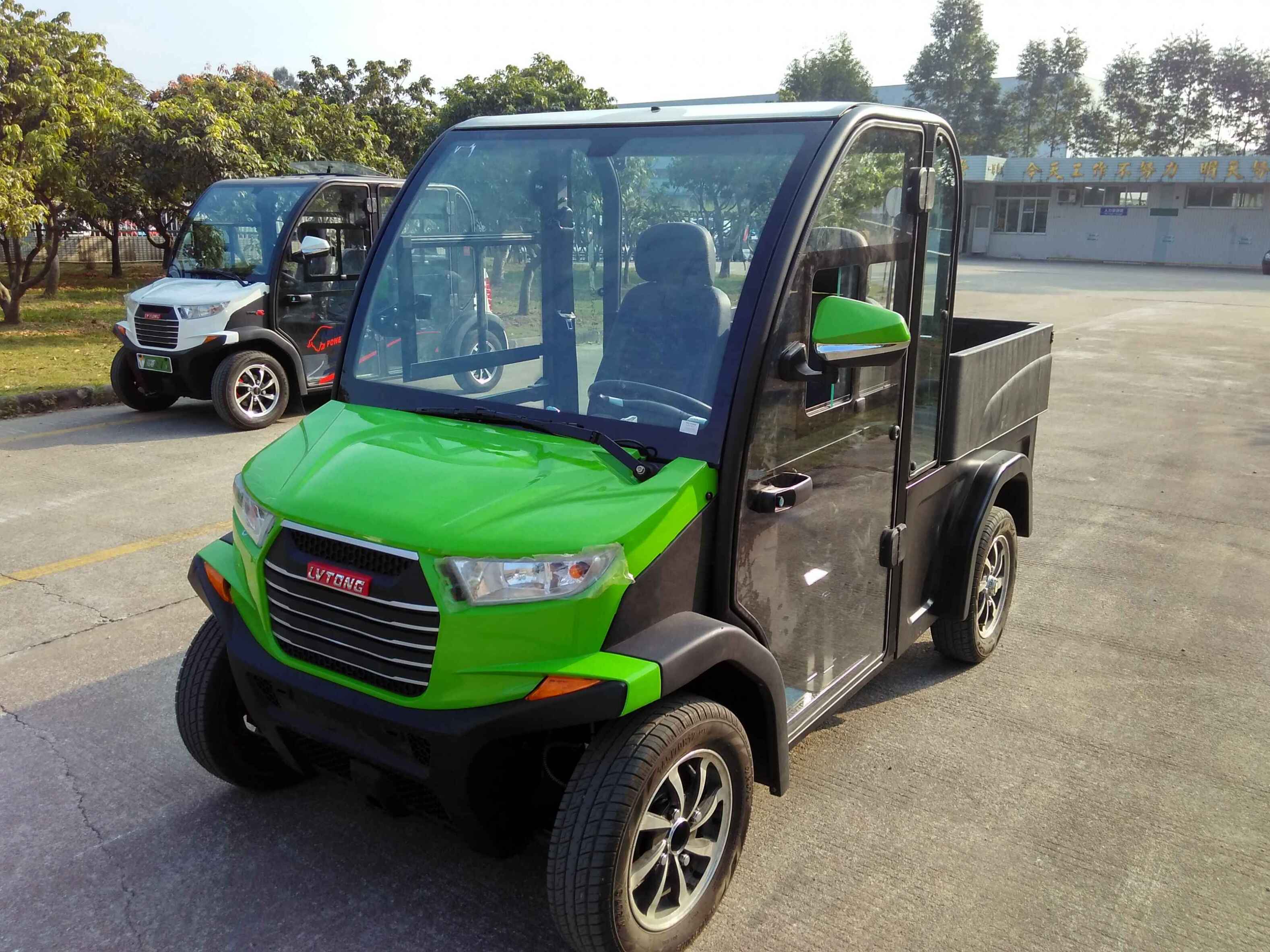 ocean cartz three-quarter front view of a lime green and black utility electric vehicle with a fully enclosed cab and a black rear cargo bed, parked on an asphalt lot. This two-seater commercial NEV or electric work cart features a bright green hood, black enclosed doors with glass, and a modern front grille. Another similar vehicle is parked in the background, suggesting a fleet. The utility transport vehicle is outdoors with trees and buildings in the distance.