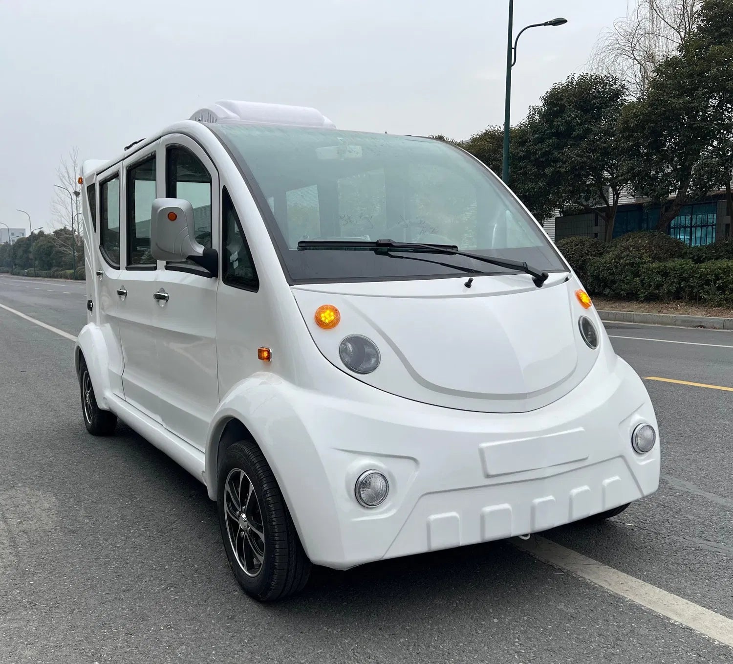 Small white electric vehicle on a road with trees and buildings in the background