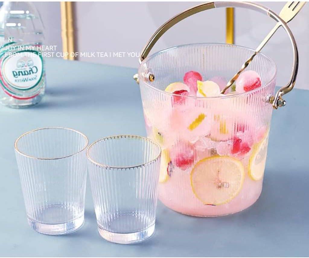 Clear glass bucket with pink lemonade and ice cubes, accompanied by two clear glasses on a light blue surface.